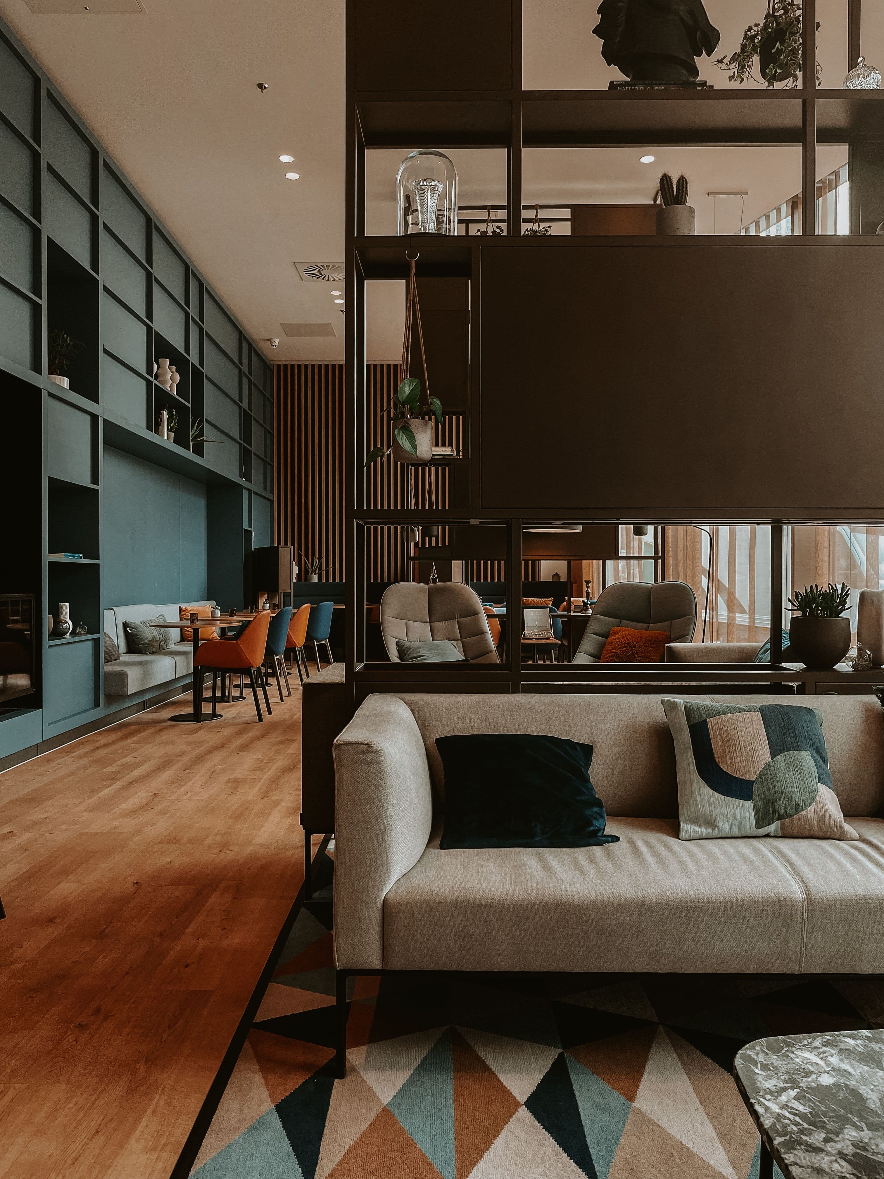 Un lobby d'hôtel contemporain aux tons bleu canard et terracotta, où canapé, banquettes et tables de travail s'organisent autour d'une claustra métallique noire, sous un plafond de grande hauteur.